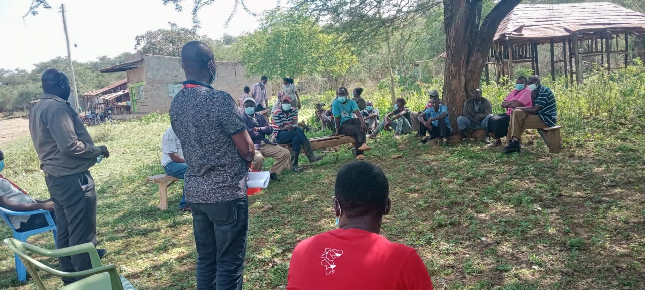 Group of people sitting under a tree listening to two people standing infront of them. Some houses and green landscape in the background