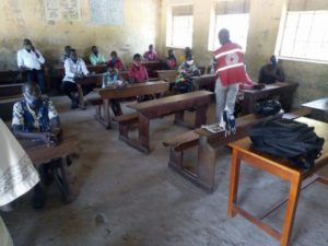 group of pupils in a classroom listening to a person in Red Cross branded vest; wall and 3 windows in the background