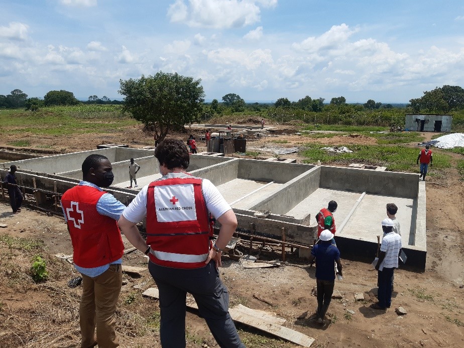 2 men standing close to the camera with their backs to the photo. Photo shows a construction site and green landscape as well as 4 other men talking right at the construction site and one person standing inside the construction site.