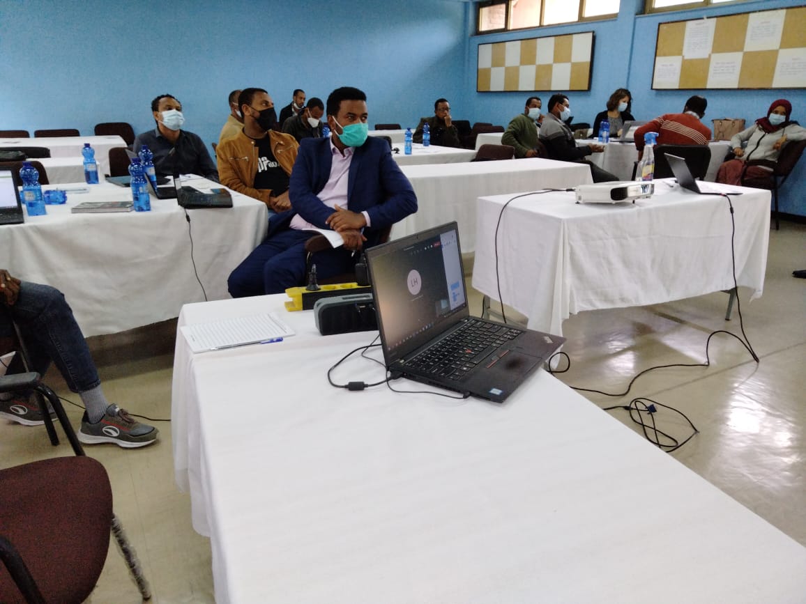 Around 15 people are sitting around tables with physical distance and masks. Some have laptops infront of them. Everyone is looking at the front of the room where the presenetation screen is hanging on the wall.