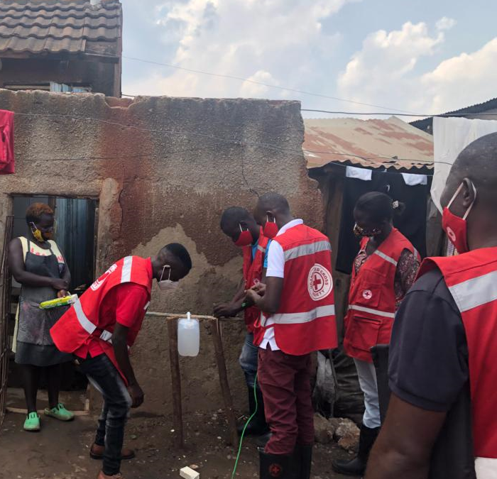 In the background a woman wearing a mask stands in the door to her house holding a soap bar. She watches 4 Red Cross volunteers wearing masks installing a tippy tap for handwashing