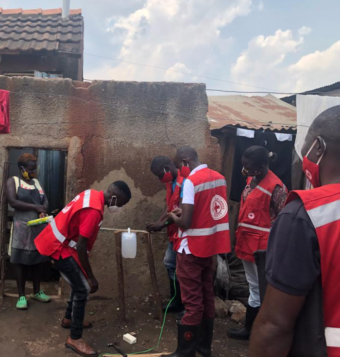 In the background a woman wearing a mask stands in the door to her house holding a soap bar. She watches 4 Red Cross volunteers wearing masks installing a tippy tap for handwashing
