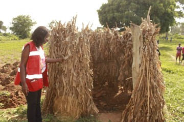 Woman in Red Cross vest inspecting a latrine made up of reeds, without a door and roof; trees and open grass in the background